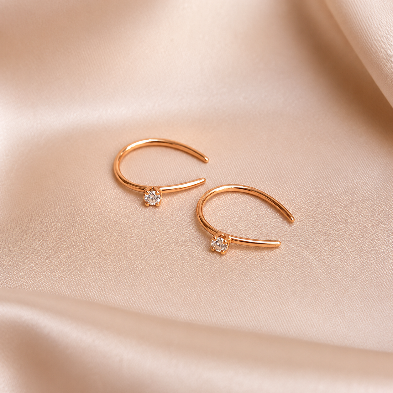 A pair of delicate curved gold-toned hoop earrings with a small clear gemstone at the center, displayed on a draped beige satin fabric background.